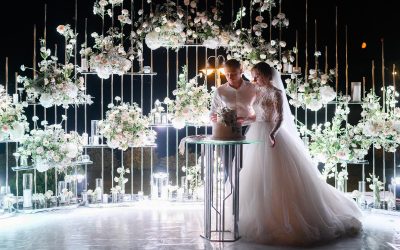 Side view of loving brides couple cutting flowering white cake on evening wedding ceremony, woman wearing in beautiful puffy dress and man in white shirt,standing near lighting floral arch,