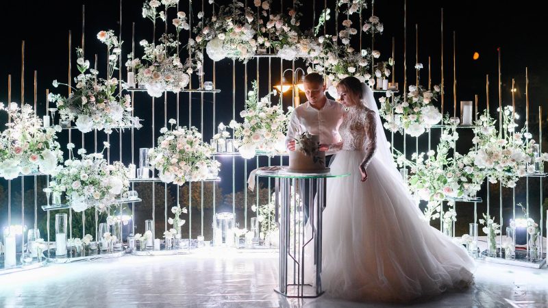 Side view of loving brides couple cutting flowering white cake on evening wedding ceremony, woman wearing in beautiful puffy dress and man in white shirt,standing near lighting floral arch,