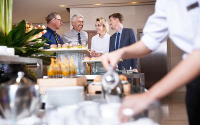 Distant four smiling business people chatting and standing at buffet table with food and blurred crockery and waiter in foreground