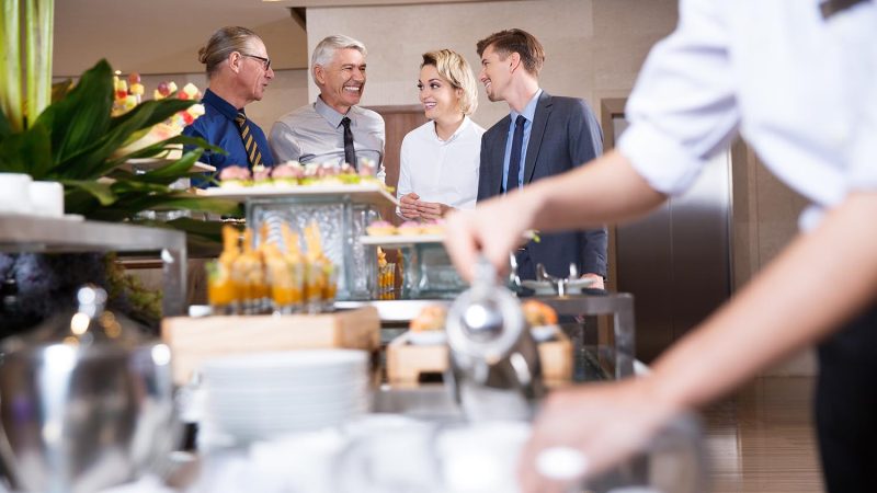 Distant four smiling business people chatting and standing at buffet table with food and blurred crockery and waiter in foreground